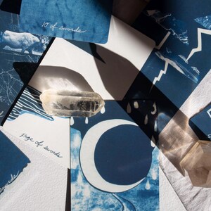 May include: A collection of indigo blue tarot cards with white illustrations and handwritten text. The cards feature images of a crescent moon, a road, and other celestial and natural elements. A clear quartz crystal is placed on top of the cards.
