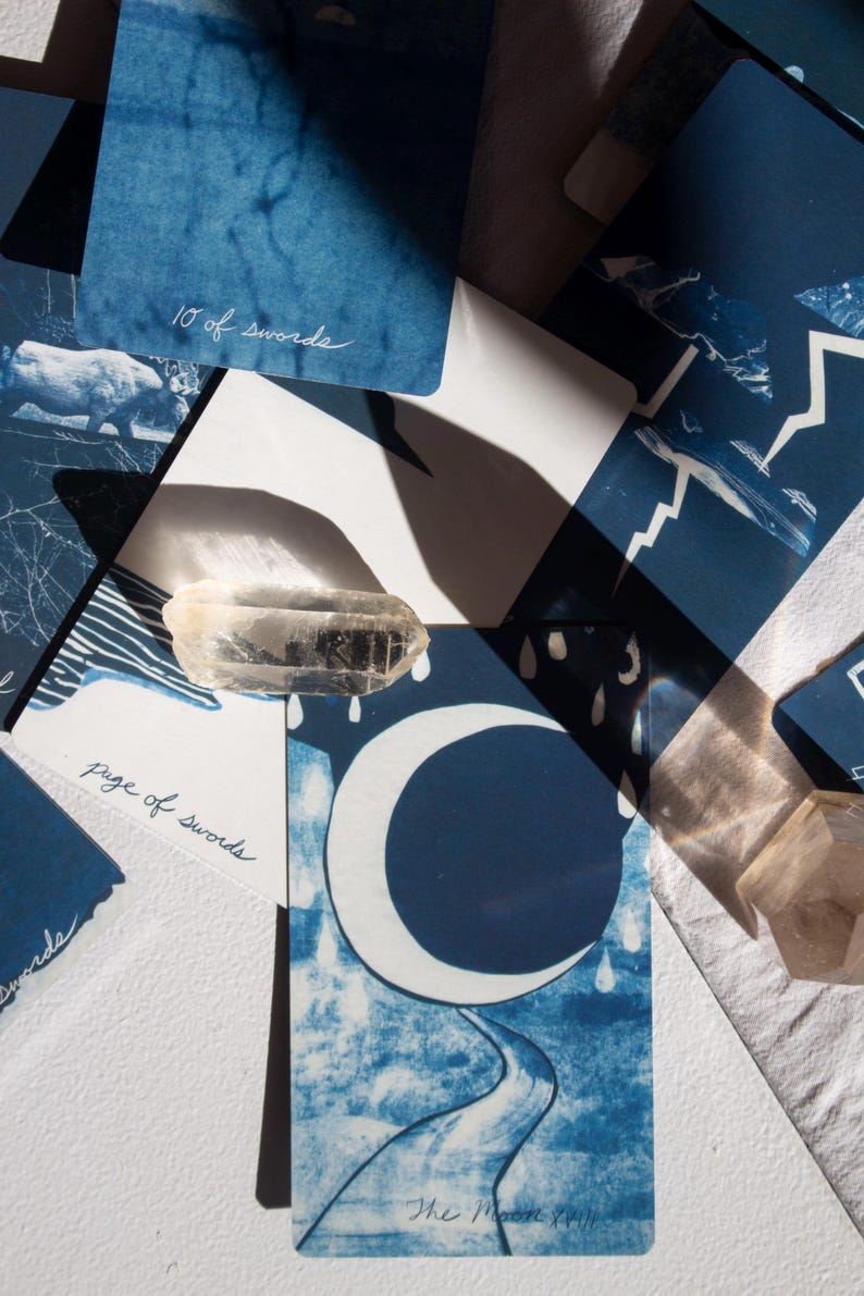 May include: A collection of indigo blue tarot cards with white illustrations and handwritten text. The cards feature images of a crescent moon, a road, and other celestial and natural elements. A clear quartz crystal is placed on top of the cards.