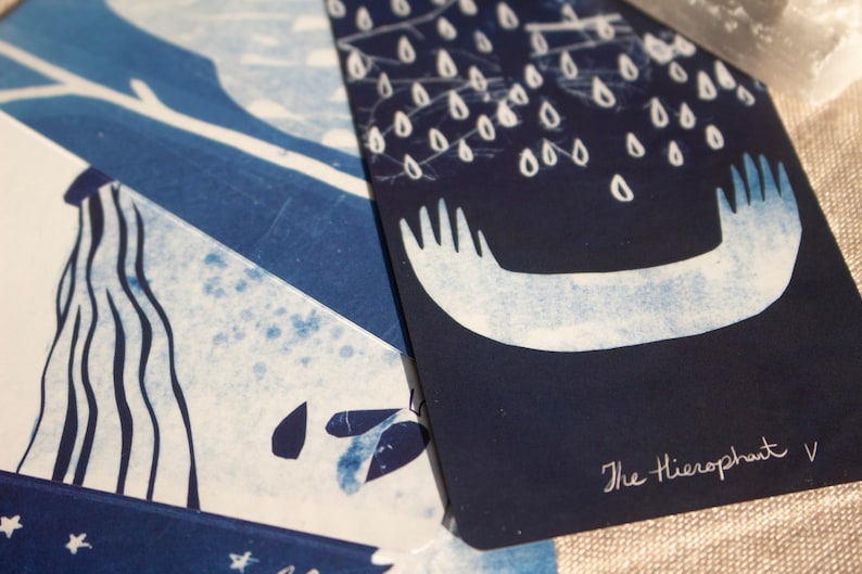 May include: Two tarot cards with indigo and white illustrations. One card features a waterfall design, and the other depicts a stylized image of hands with raindrops. The text "The Hierophant V" is visible on the second card.
