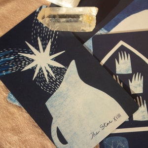 May include: A close-up of tarot cards with celestial and hand-drawn imagery. The cards are primarily navy blue with white and light blue illustrations. One card features a star, another shows hands. A clear crystal rests on the cards.