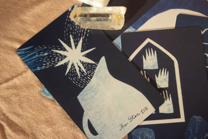 May include: A close-up of tarot cards with celestial and hand-drawn imagery. The cards are primarily navy blue with white and light blue illustrations. One card features a star, another shows hands. A clear crystal rests on the cards.