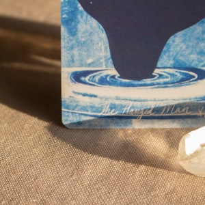 May include: A blue and white tarot card with a dark blue shape and water-like design. The text "The Hanged Man XII" is visible. A clear crystal is in the foreground, with a neutral-toned fabric background.