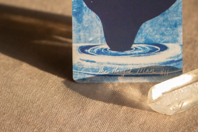 May include: A blue and white tarot card with a dark blue shape and water-like design. The text "The Hanged Man XII" is visible. A clear crystal is in the foreground, with a neutral-toned fabric background.