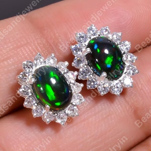 Puede incluir: Un par de pendientes de plata con piedras de ópalo negro y verde rodeadas por un halo de pequeñas piedras de circonita cúbica transparentes.