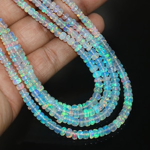 Puede incluir: Varias hebras de collares de cuentas de ópalo. Las cuentas son pequeñas, redondas e iridiscentes, mostrando colores como el azul, el verde y el rosa. Los collares se presentan sobre un fondo oscuro.