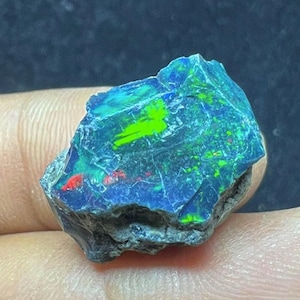 Op de afbeelding: Een ruwe opaal edelsteen met een donkere basiskleur, met levendige flitsen van blauw, groen en rood. De steen heeft een getextureerd oppervlak en een onregelmatige vorm, met natuurlijke patronen en kleuren.