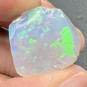 Op de afbeelding: Een ruwe, ongeslepen opaal edelsteen met een melkachtige witte basiskleur. De steen vertoont levendige flitsen van groen, blauw en hints van rood over het oppervlak. De opaal heeft een ruwe, natuurlijke textuur en onregelmatige vorm.