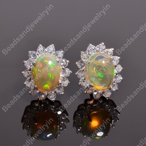May include: A pair of opal stud earrings. Each earring features an oval-shaped opal gemstone surrounded by a halo of small, clear gemstones. The opals display a play of color with flashes of green, yellow, and orange. The earrings are set on a reflective surface.
