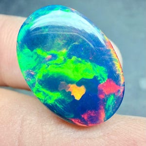 May include: An oval-shaped black opal gemstone featuring a dark blue base color with vibrant, multi-colored flashes of neon green, bright orange, and red throughout the surface, displaying a polished, smooth, and reflective finish.