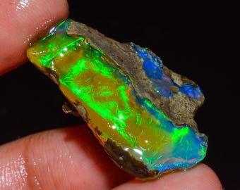May include: A vibrant, raw opal gemstone with a striking display of color. The opal features a mix of bright green, yellow, and blue hues, with a rough, textured exterior. The gemstone is held between two fingers, showcasing its natural beauty.