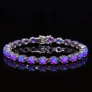 May include: A silver bracelet featuring a series of round, iridescent opal stones. The opals display a mix of blue and purple hues, set in a silver metal band. The bracelet is displayed against a black background.