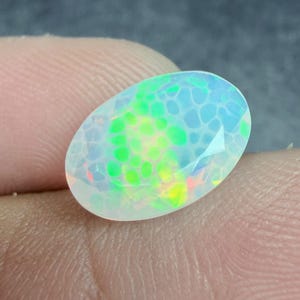 May include: An oval-shaped, faceted opal gemstone displaying a translucent white base with a vibrant play-of-color featuring bright neon green, yellow, and blue honeycomb-like patterns across its surface, reflecting light with a distinct iridescent sheen.