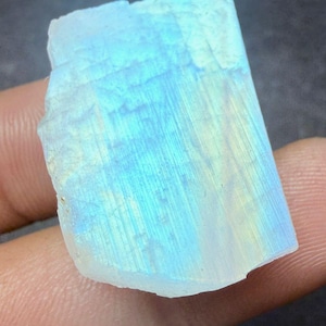 May include: A raw, rectangular moonstone gemstone with a pale blue and white color. The stone has a translucent quality, with subtle internal lines and a slightly rough texture. The gemstone is held in a hand.