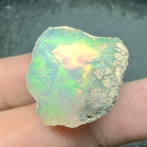Op de afbeelding: Een ruwe opaal edelsteen met een levendig kleurenspel. De steen toont flitsen van groen, geel en roze in een doorschijnende, melkachtige witte basis. De ruwe randen vertonen een getextureerd, natuurlijk uiterlijk.