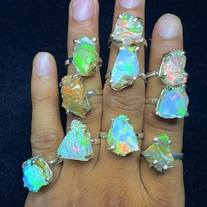 May include: A hand adorned with several silver rings, each showcasing a distinctive, raw opal stone. The opals exhibit a range of colours, including green, blue, orange, and white, with iridescent flashes. The rings are set against a dark backdrop.