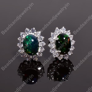 May include: A pair of silver-toned stud earrings. Each earring features a dark green, oval-shaped gemstone surrounded by a halo of smaller, clear gemstones. The earrings are set against a dark, reflective surface.