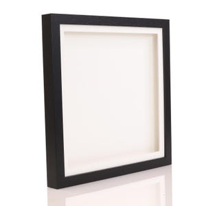 May include: A square shadow box frame with a black exterior and a white interior. The frame is made of wood and has a deep profile, creating a three-dimensional effect. The frame is designed to display artwork or other items.