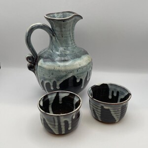 May include: A ceramic pitcher with a curved handle and two matching small cups, all featuring a dark brown base glaze with a contrasting white drip pattern that cascades down from the rim, creating a textured and layered appearance on each piece.