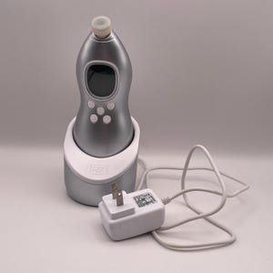 May include: A silver-colored skincare device with a white base and a charging adapter. The device has a digital display and several buttons. The adapter has a two-prong plug and a white cord. The device is designed for skincare treatments.