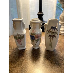 May include: Three small white ceramic vases arranged in a row. The left vase features a painted cluster of pink grapes with green leaves. The center vase displays a floral pattern with blue and pink petals. The right vase shows a green bamboo design.