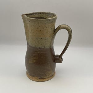 May include: A ceramic pitcher featuring a two-tone glaze design. The upper portion is a speckled light beige, while the lower body is a solid dark brown. It includes a curved handle attached to the side and a small spout at the top rim.