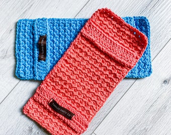 May include: Two handmade crochet phone cases in bright colors. One is a vibrant coral, the other is a light blue. Each case has a rectangular shape with a textured stitch pattern and a small, dark brown label that reads "Tangled Knots Co."