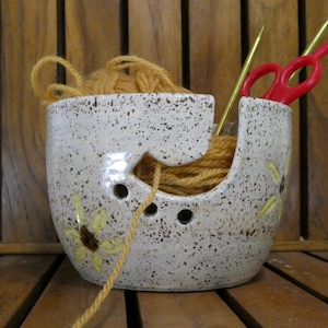 May include: A speckled ceramic yarn bowl with a sunflower design. The bowl holds yellow yarn, knitting needles, and red scissors. The bowl has a curved opening for the yarn to feed through and three small holes.