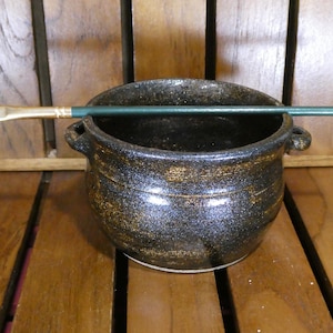 May include: A small, round, dark ceramic bowl with two handles and a gold speckled finish. A green-handled paintbrush with a white brush head rests across the top of the bowl. The bowl sits on a wooden surface.