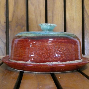 May include: A handmade ceramic butter dish with a lid. The dish is a deep red with a speckled texture, and the lid has a turquoise knob. The dish sits on a matching red plate with a cream-colored rim. The pottery is glazed and appears glossy.