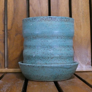 May include: A turquoise ceramic planter with a matching saucer. The planter has a textured, ridged design and is speckled with dark brown spots. The handmade pottery is displayed on a wooden surface.
