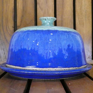 May include: A ceramic butter dish with a domed lid and a matching base. The dish is glazed in shades of blue, transitioning from a light teal at the top to a deep cobalt blue at the base. The dish sits on a wooden surface.