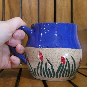 May include: A handmade ceramic mug with a blue glazed top and a beige speckled bottom. The mug features a painted design of red flowers and green leaves. The handle is blue.