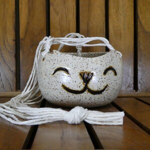 May include: A speckled ceramic cat-shaped hanging planter with a smiling face design. The planter is off-white with brown speckles and a brown nose and mouth. It hangs from a white macrame cord, perfect for small plants.