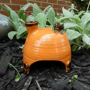 May include: A bright orange ceramic toad house with a glossy finish, featuring a small brown chimney on the top left and a sculpted green frog figurine perched on the right side. The structure has a wide arched opening at the base for entry.