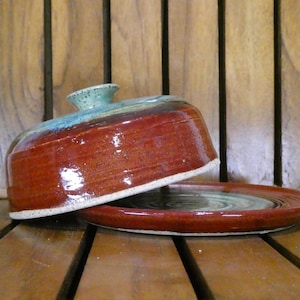 May include: A handmade ceramic butter dish with a lid. The dish is a deep red-brown color with a turquoise and green rim and knob. The dish is sitting on a wooden surface.