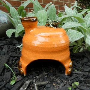 May include: A bright orange ceramic toad house with a glossy finish, featuring a small sculpted green frog perched on the top and a dark brown chimney-like opening on the left side, supported by four rounded brown feet at the base.