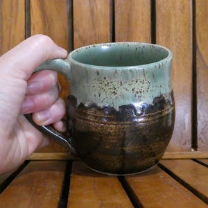May include: A handmade ceramic mug with a green and brown glaze. The mug has a rounded shape with a handle and is held by a hand. The mug's top is green, transitioning to a speckled tan and then a dark brown.