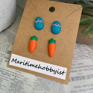 Peut inclure: Un ensemble de quatre boucles d'oreilles en pâte polymère comprenant deux motifs d'œufs de Pâques bleus et roses et deux motifs de carottes orange. Les boucles d'oreilles sont présentées sur une carte brune avec le texte "Maritimehobbyist".