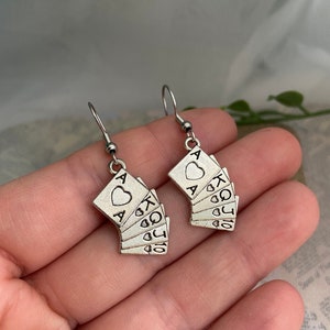 May include: A pair of silver earrings shaped like a fan of playing cards. The cards are all hearts and include the Ace, King, Queen, Jack, 10, 9, and 8.
