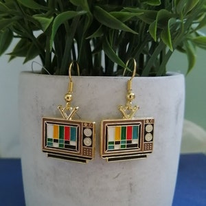 May include: Gold-tone earrings featuring a retro television design with a colourful test pattern on the screen. The earrings have a brown and black finish.