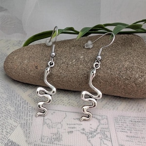 May include: A pair of silver-toned metal earrings featuring a dangling snake charm. Each earring has a curved, S-shaped body with a smooth finish and a simple hook-style ear wire with a small, clear plastic backing for secure placement.