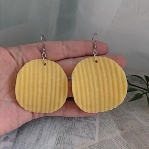 May include: A pair of novelty earrings shaped like yellow, ridged potato chips. The earrings are suspended from silver-colored fish hook ear wires. The earrings are a fun, quirky accessory.