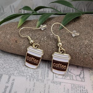 May include: A pair of gold-tone earrings with a coffee cup charm. Each charm is white with a brown coffee cup design and the word "coffee" printed on it.