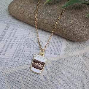 May include: A gold-coloured chain necklace featuring a pendant shaped like a coffee cup. The pendant is white with a brown band and the word "coffee" in white. The necklace is displayed on a stone.