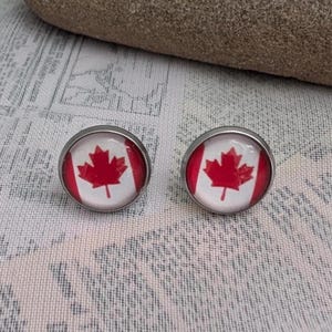 May include: A pair of round stud earrings featuring the Canadian flag design with a red maple leaf centred on a white background flanked by two red vertical bars, all encased in a polished silver-toned metal setting with a smooth glass dome finish.