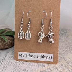 May include: Silver-coloured dangle earrings with coffee bean and espresso maker charms. The earrings are displayed on a brown card with the text "Maritime Hobbyist". The coffee bean charms have vertical lines, and the espresso maker charms have colourful accents.
