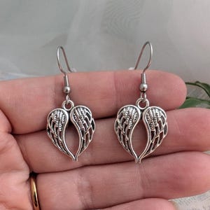 May include: A pair of silver-colored earrings. Each earring features a heart-shaped design composed of stylized wings. The earrings have a hook closure. The jewelry is displayed against a soft, neutral background.