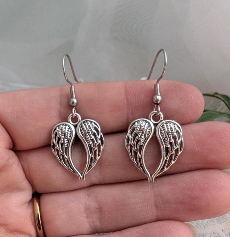 May include: A pair of silver-colored earrings. Each earring features a heart-shaped design composed of stylized wings. The earrings have a hook closure. The jewelry is displayed against a soft, neutral background.