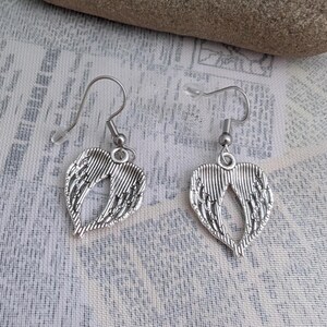 May include: A pair of silver-colored earrings featuring a heart-shaped wing design. The earrings have a hook closure and are displayed on a patterned fabric background. The wings have detailed feather-like lines.