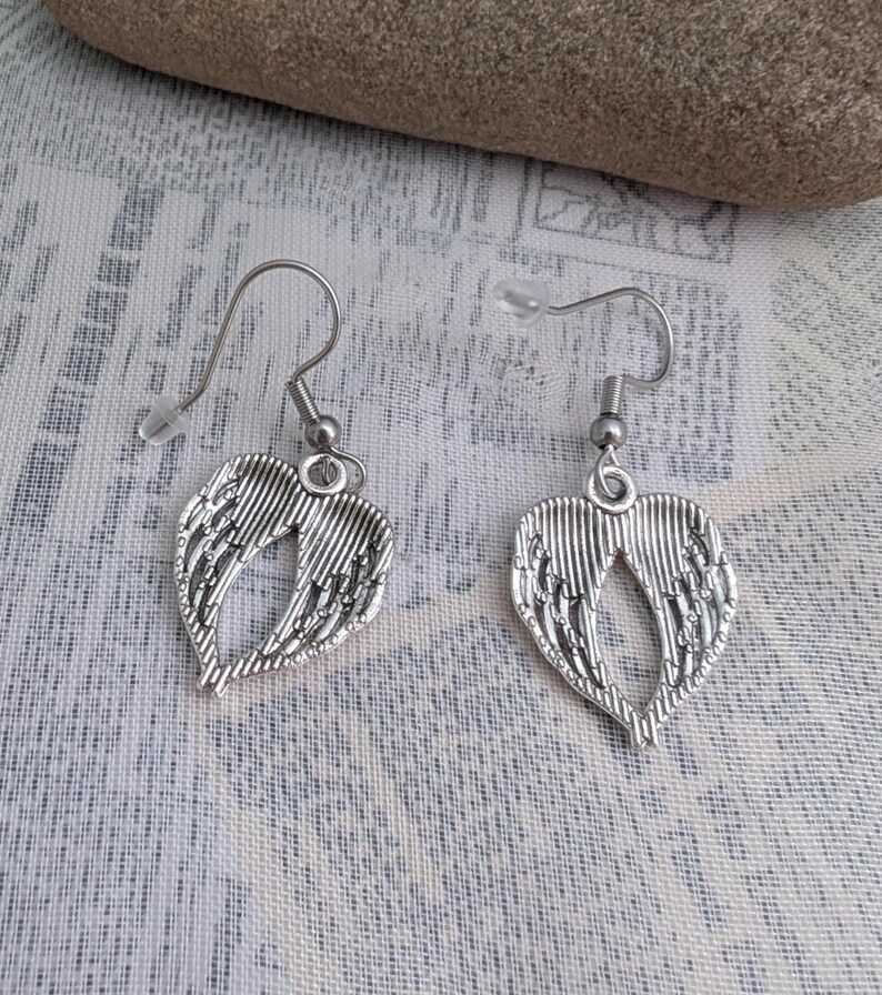 May include: A pair of silver-colored earrings featuring a heart-shaped wing design. The earrings have a hook closure and are displayed on a patterned fabric background. The wings have detailed feather-like lines.